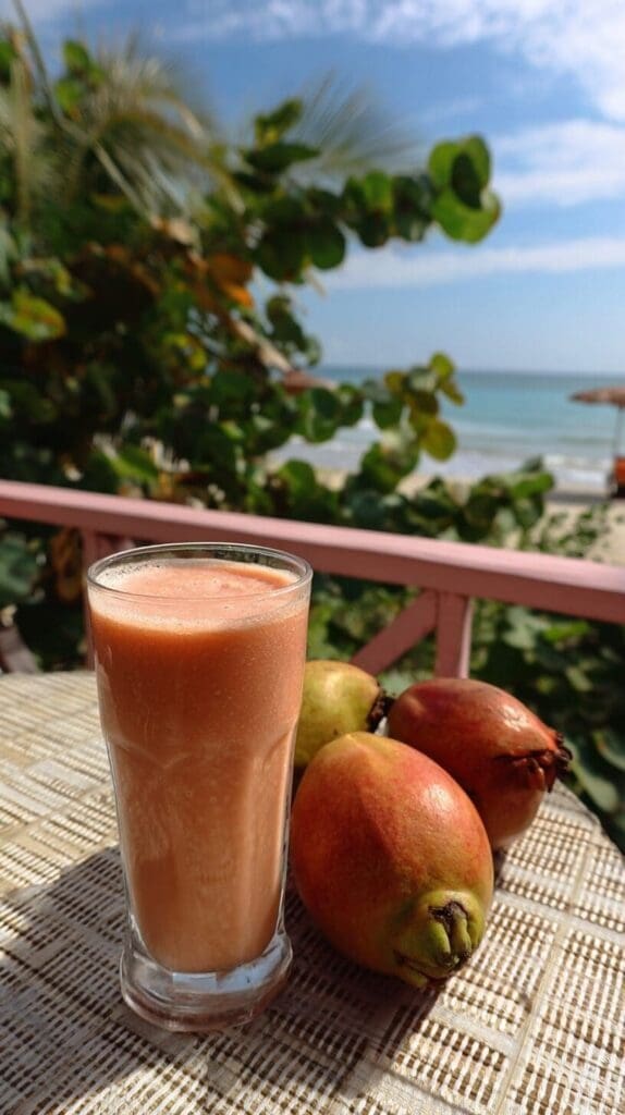A glass of light orange-pink smoothie sits on a patterned outdoor table next to two fresh guavas. In the background, a sunny beach and ocean are visible through lush green foliage.